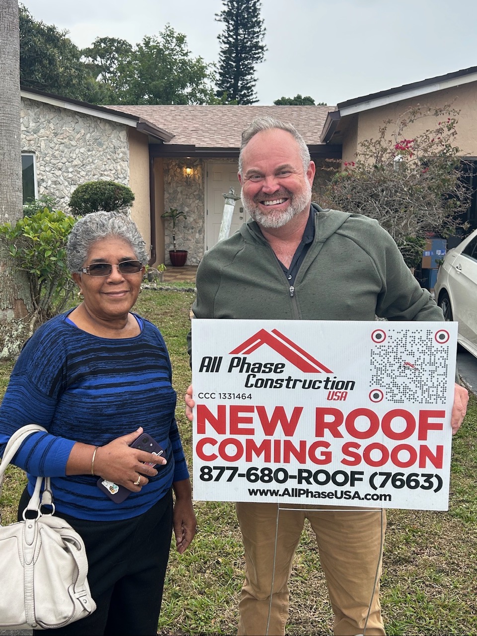 All Phase Construction representative Graham with happy customer holding new roof sign in South Florida
